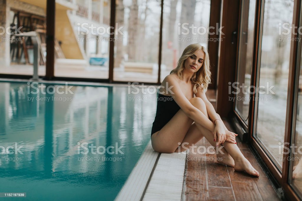 Attractive young blonde woman in bikini and with sunglasses sits by the poolside in the indoor swimming pool
